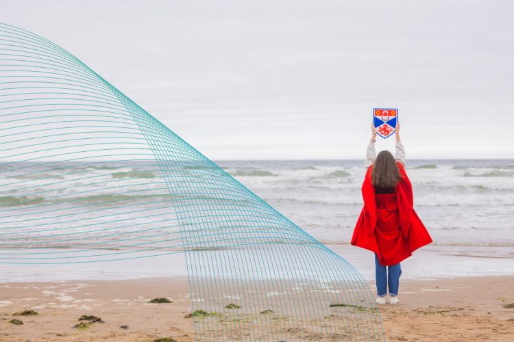 A female student in a red gown looks out towards the sea, holding a University of St Andrews crest above her head.